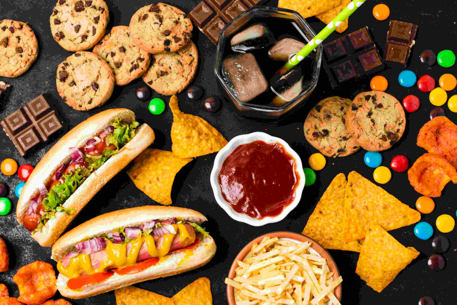 Ultra-processed food including hot dogs, cookies, chips, and soda on a dark background.